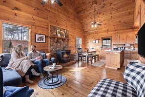 A cozy cabin interior with a fireplace, wooden walls, a dining area, and a kitchen.