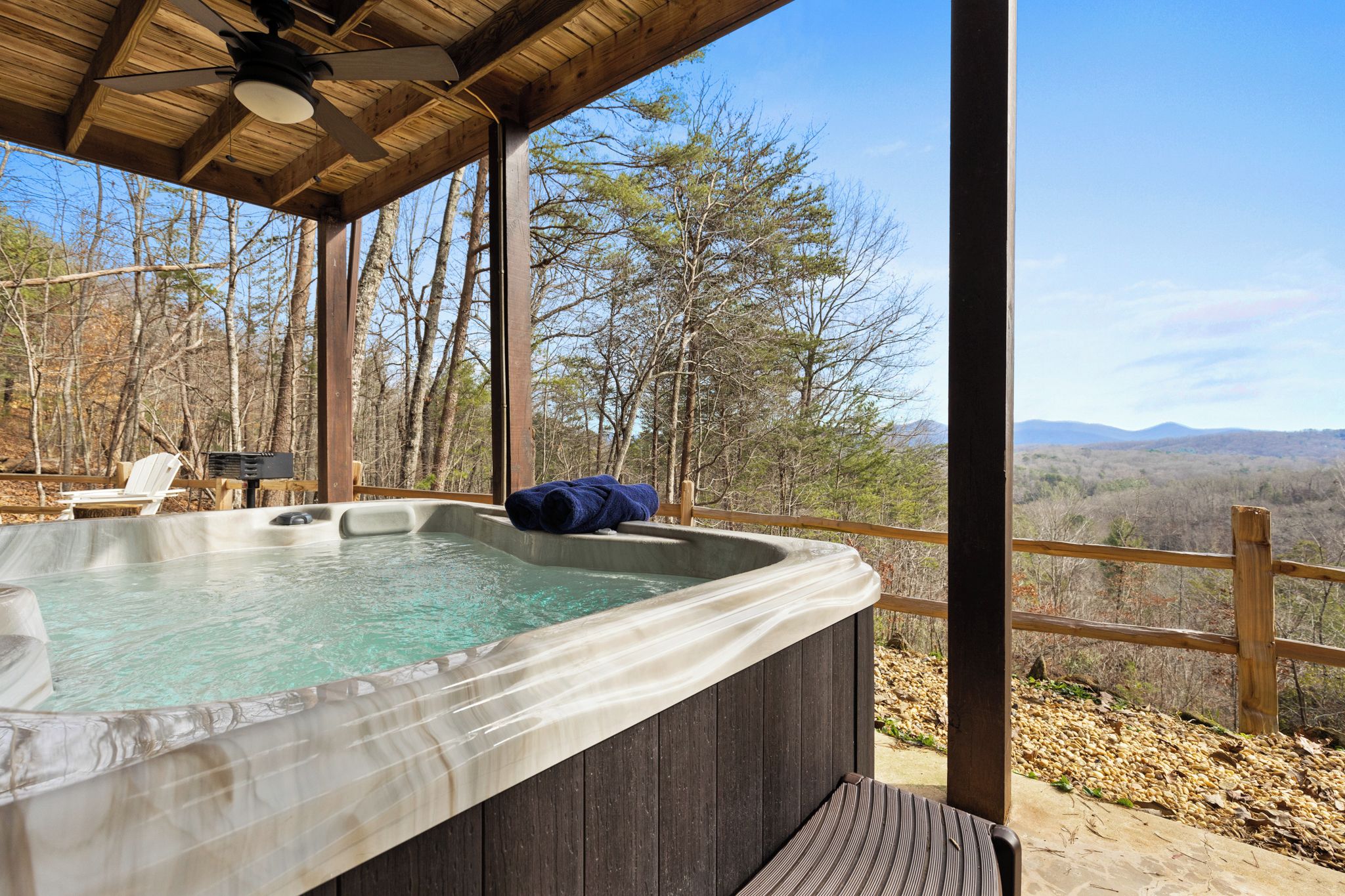 A hot tub with a wooden deck and a wooden railing, surrounded by trees and a mountainous landscape.