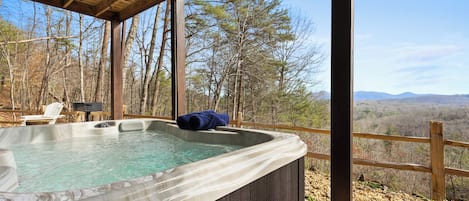 A hot tub with a wooden deck and a wooden railing, surrounded by trees and a mountainous landscape.