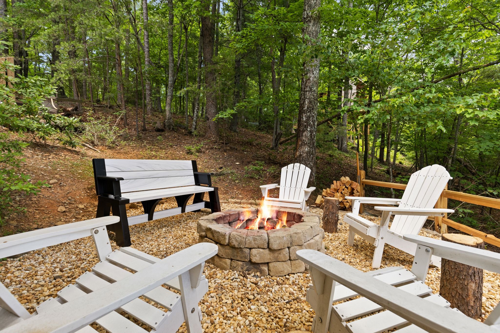 A fire pit surrounded by Adirondack chairs and a bench in a forested area.
