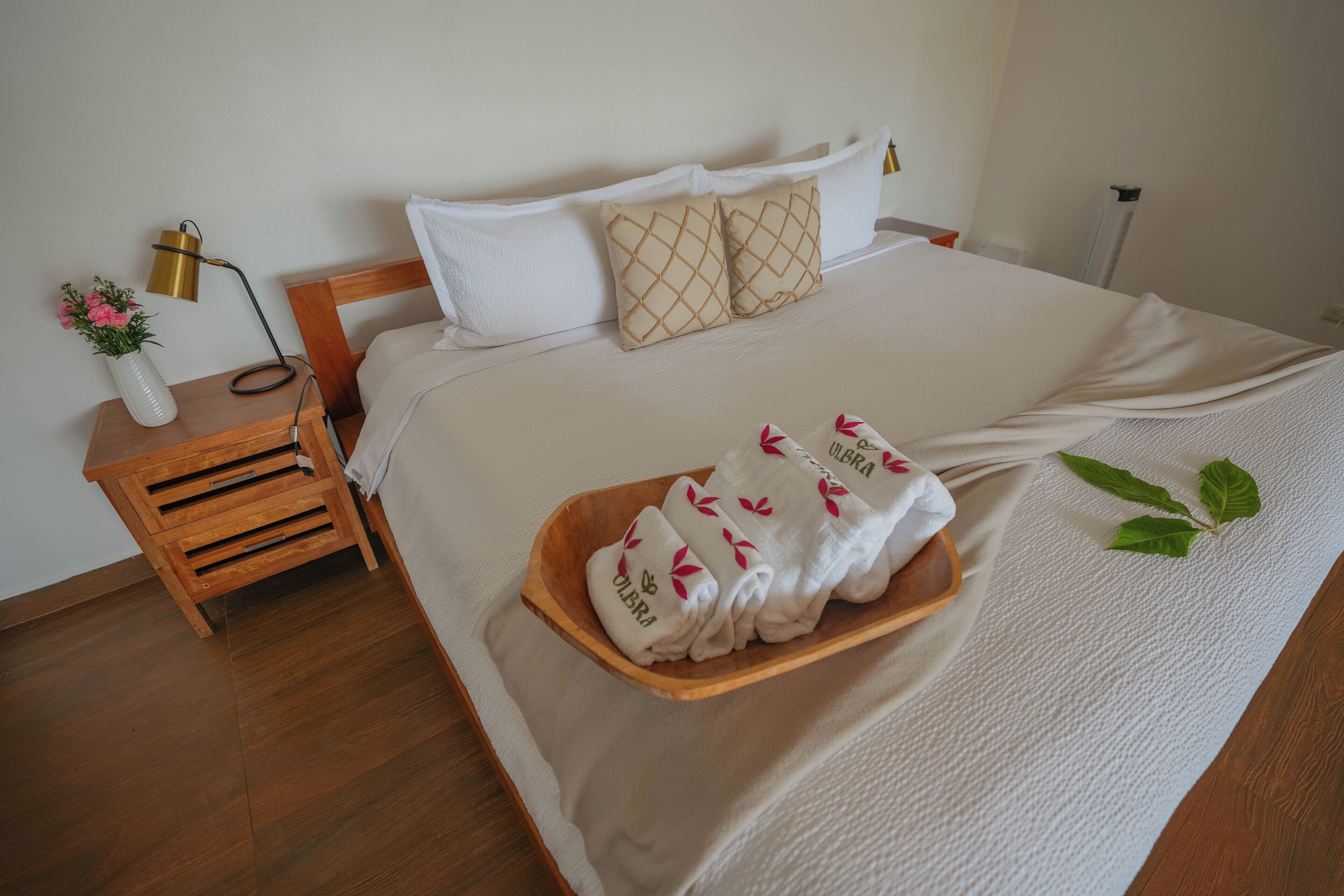 Una cama bien tendida con ropa blanca, una mesita de noche de madera con una lámpara, y una bandeja con toallas dobladas.
