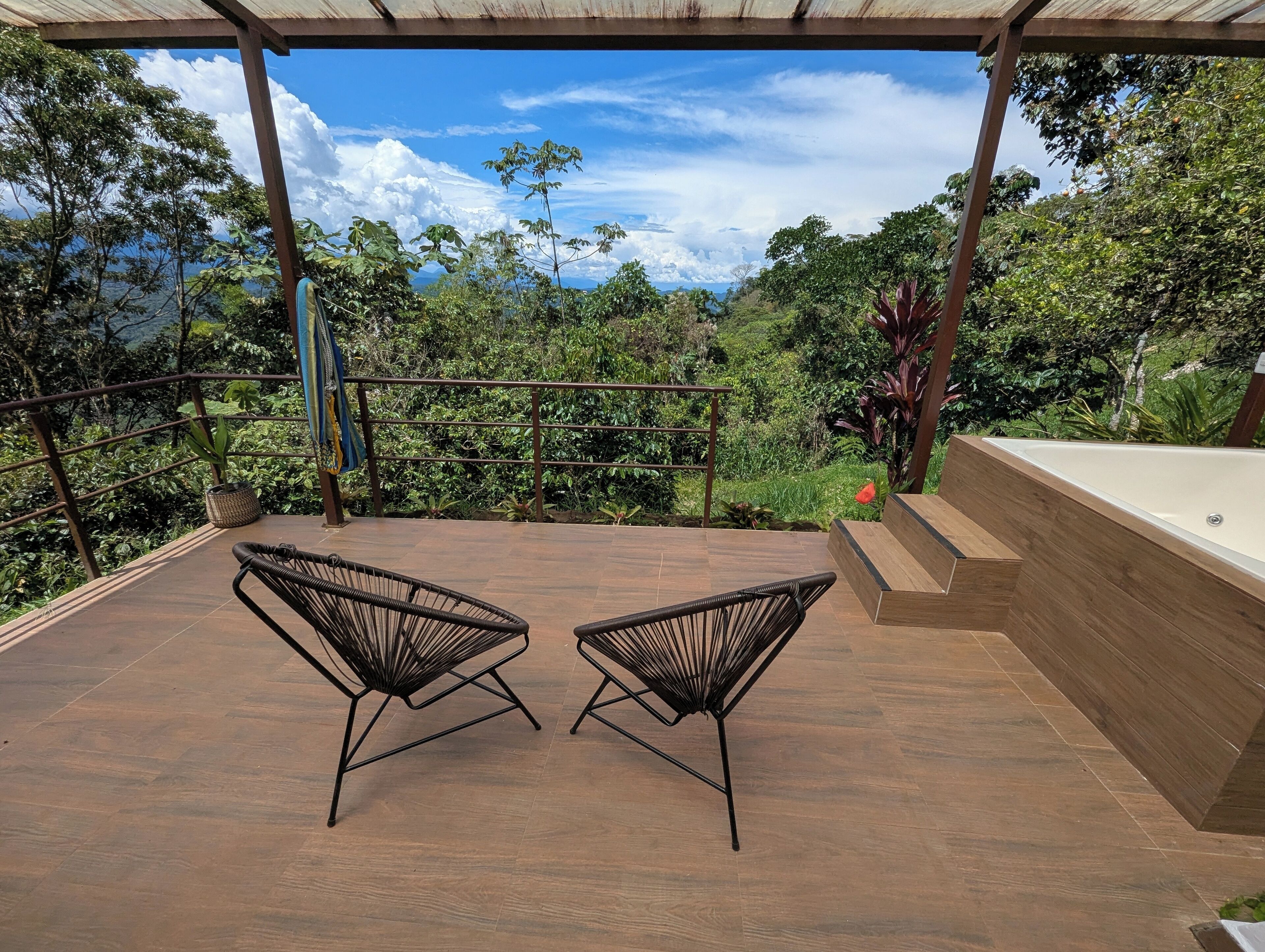 An outdoor patio with two black wireframe chairs, a wooden deck, and a view of a lush green landscape and the sea.