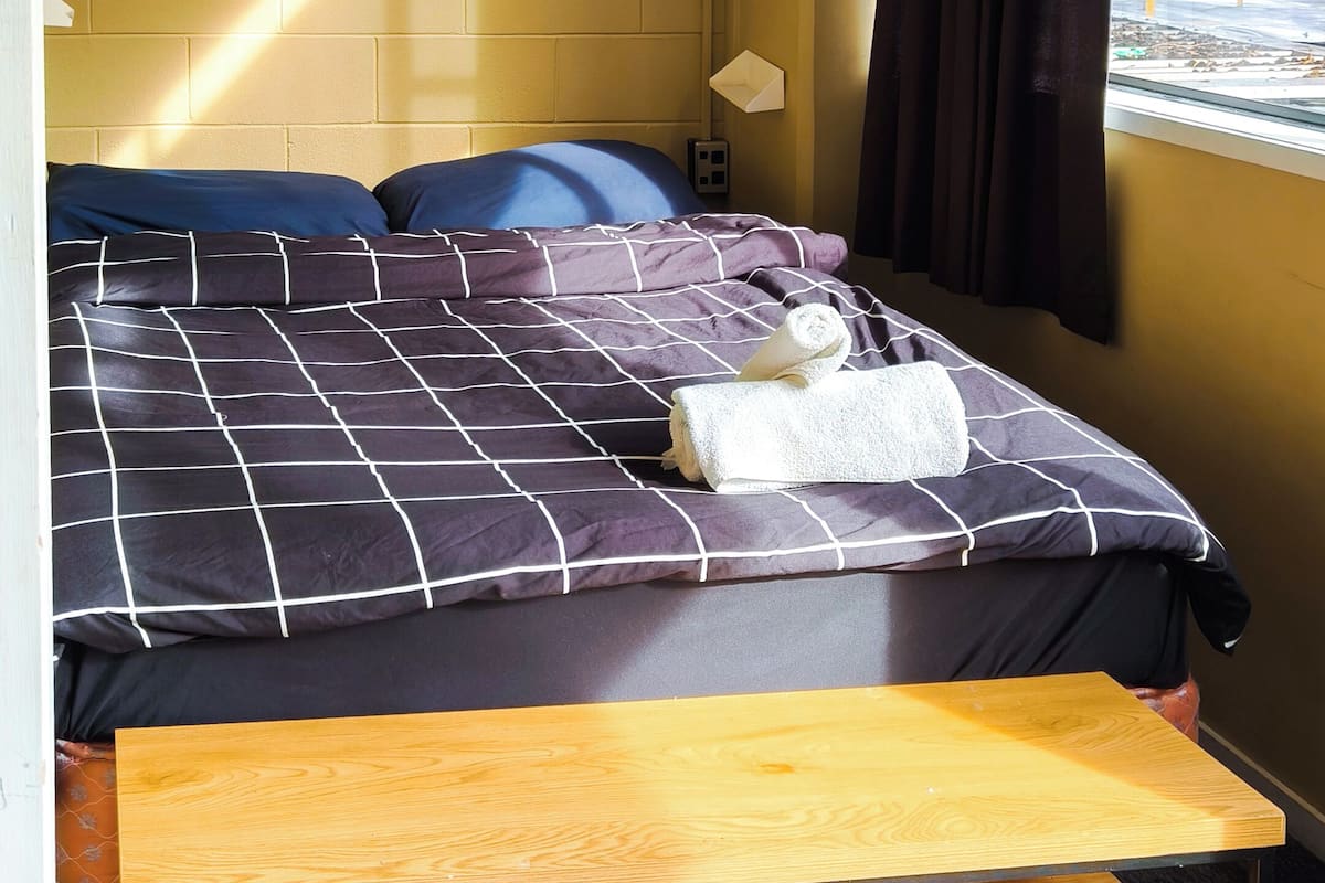 A hotel room with a bed, a wooden bench, a window with curtains, and a wall with a brick pattern.