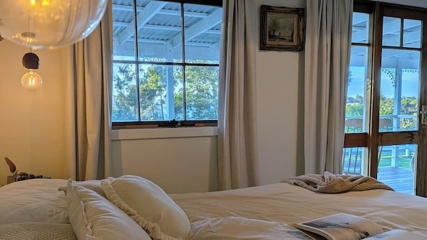A bedroom with a large bed, a window with curtains, a hanging light fixture, and a framed picture on the wall. - Cute River Shack with Wood Fire in Clarence Point. (Clarence Point)