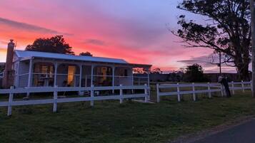 A rustic building with a porch, surrounded by a white fence, set against a sunset sky.