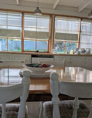 A kitchen with a wooden table, white chairs, a bowl of fruit, and a window with curtains.