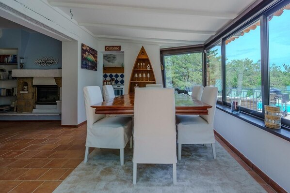 A dining area with a wooden table and white chairs, a kitchen in the background, and a view of trees through large windows.