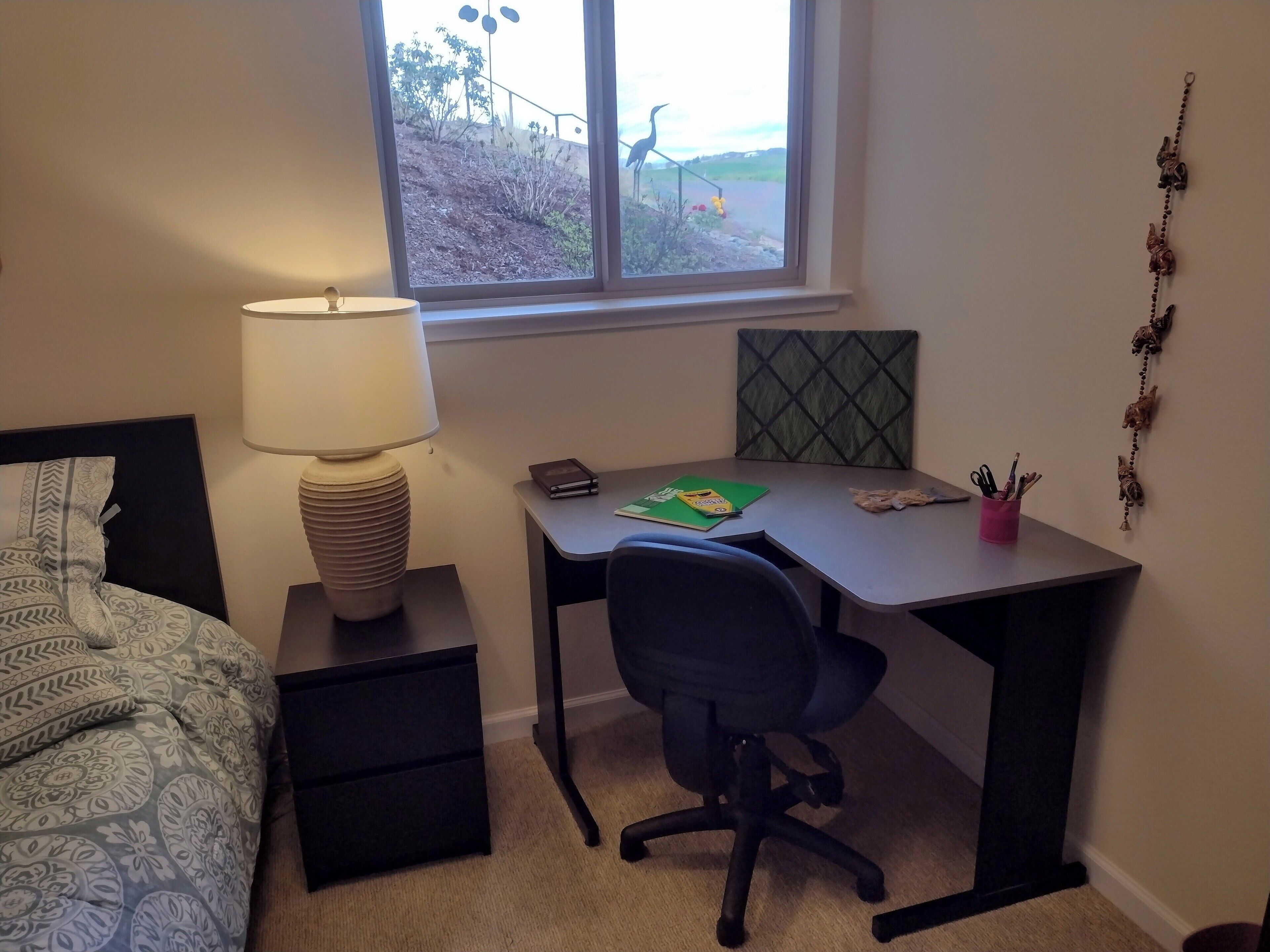 A bedroom with a bed, a desk, a chair, a lamp, and a window with a view of a garden.