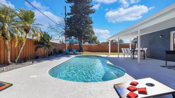 Une piscine rectangulaire à bord incurvé, entourée d’une terrasse en béton. On y trouve des chaises longues et un patio couvert avec des tables et des chaises.