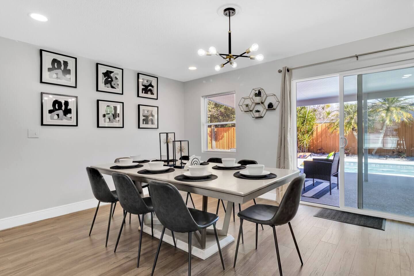 Une salle à manger avec une longue table dressée pour six personnes, entourée de chaises noires. Des œuvres d’art encadrées ornent le mur, ainsi qu’un luminaire hexagonal et une fenêtre donnant sur un jardin.