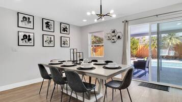 Une salle à manger avec une longue table dressée pour six personnes, entourée de chaises noires. Des œuvres d’art encadrées ornent le mur, ainsi qu’un luminaire hexagonal et une fenêtre donnant sur un jardin.