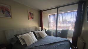 A hotel room with a large bed, a nightstand, a window with blinds, and a view of the outside. - The best location in Santiago, in the heart of the golf neighborhood, MUT, Costanera Ctr. (Las Condes)