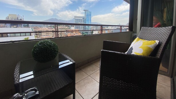 A balcony with a wicker chair and a small table, a view of a cityscape with buildings and a mountain in the distance. - The best location in Santiago, in the heart of the golf neighborhood, MUT, Costanera Ctr. (Las Condes)