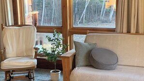 A cozy living room with a wooden rocking chair, a beige sofa, and a large window with curtains. - Private, secluded 2-bedroom house with King, Queen beds near Cable with WiFi, AC (Cable)