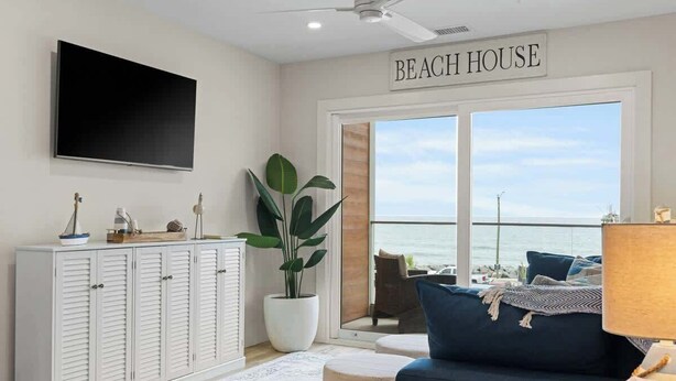 A room with a large TV, a cabinet, a plant, a sofa, and a view of the ocean. - Beach, You Crazy! by Carolina Getaway- Oceanfront 2BR Condo POOL & HOT TUB OPEN (Carolina Beach)