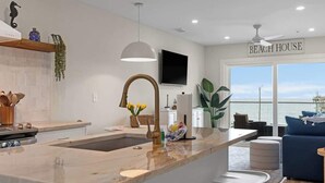 A modern kitchen with a marble island, a large window with a view, and a 'BEACH HOUSE' sign. - Beach, You Crazy! by Carolina Getaway- Oceanfront 2BR Condo POOL & HOT TUB OPEN (Carolina Beach)