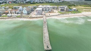 An aerial view of a coastal area with a long pier extending into the water, surrounded by residential buildings. - Beach, You Crazy! by Carolina Getaway- Oceanfront 2BR Condo POOL & HOT TUB OPEN (Carolina Beach)