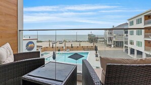 A balcony with wicker furniture, a pool, and a view of the beach and ocean. - Beach, You Crazy! by Carolina Getaway- Oceanfront 2BR Condo POOL & HOT TUB OPEN (Carolina Beach)