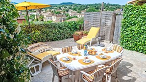 Une terrasse extérieure avec une table dressée pour le repas, entourée de chaises, et un parasol jaune offrant de l’ombre.