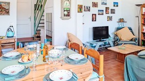 Un coin repas avec une table en bois, dressée pour quatre personnes, entourée de chaises. Un téléviseur est fixé au mur, et une bibliothèque est visible au fond.