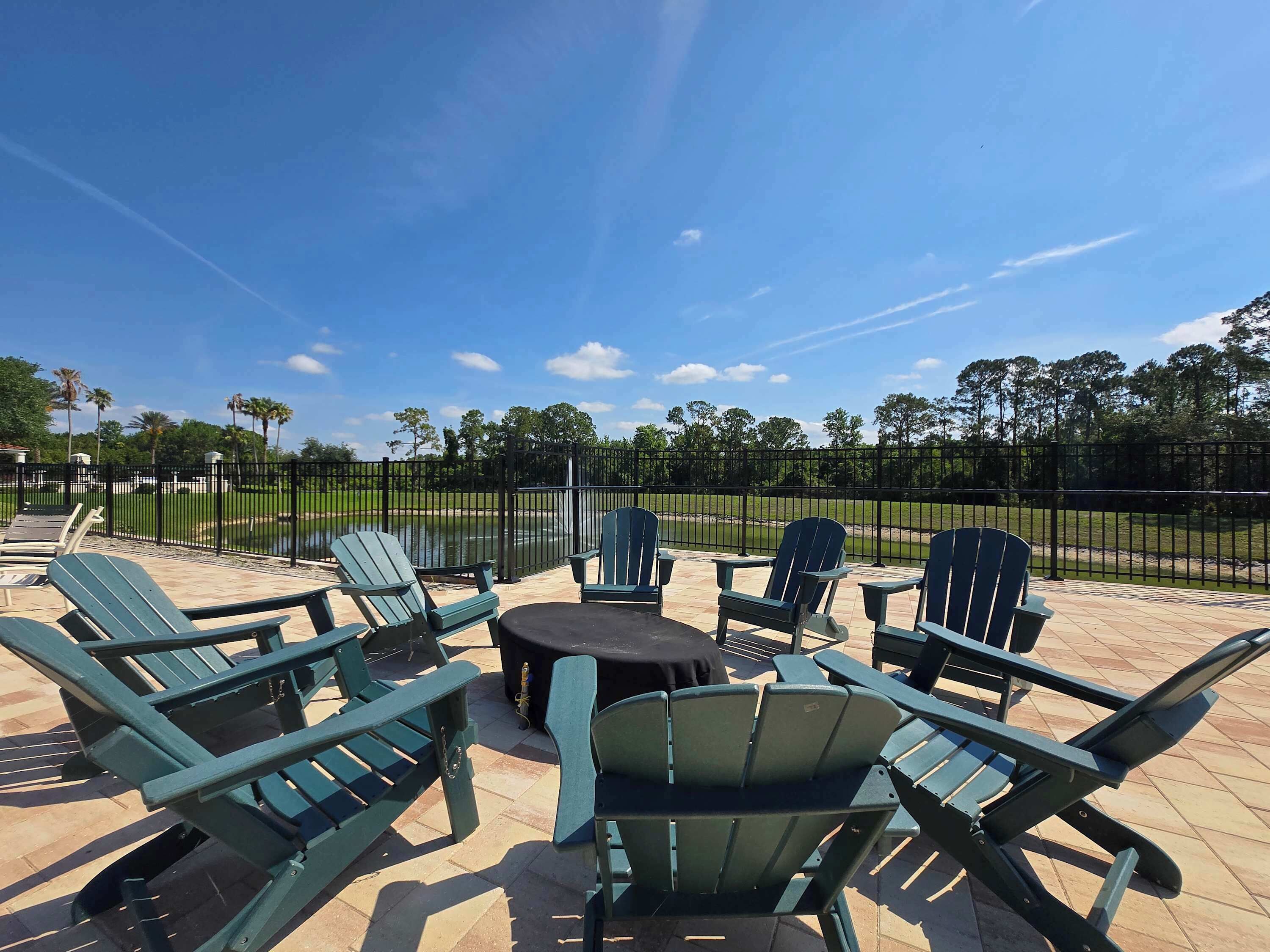 An outdoor seating area with green Adirondack chairs and a round table, surrounded by a black metal fence.