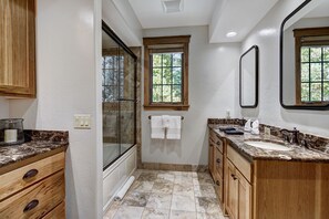 A bathroom with a glass shower enclosure, wooden cabinets, a marble countertop, and a window providing natural light.
