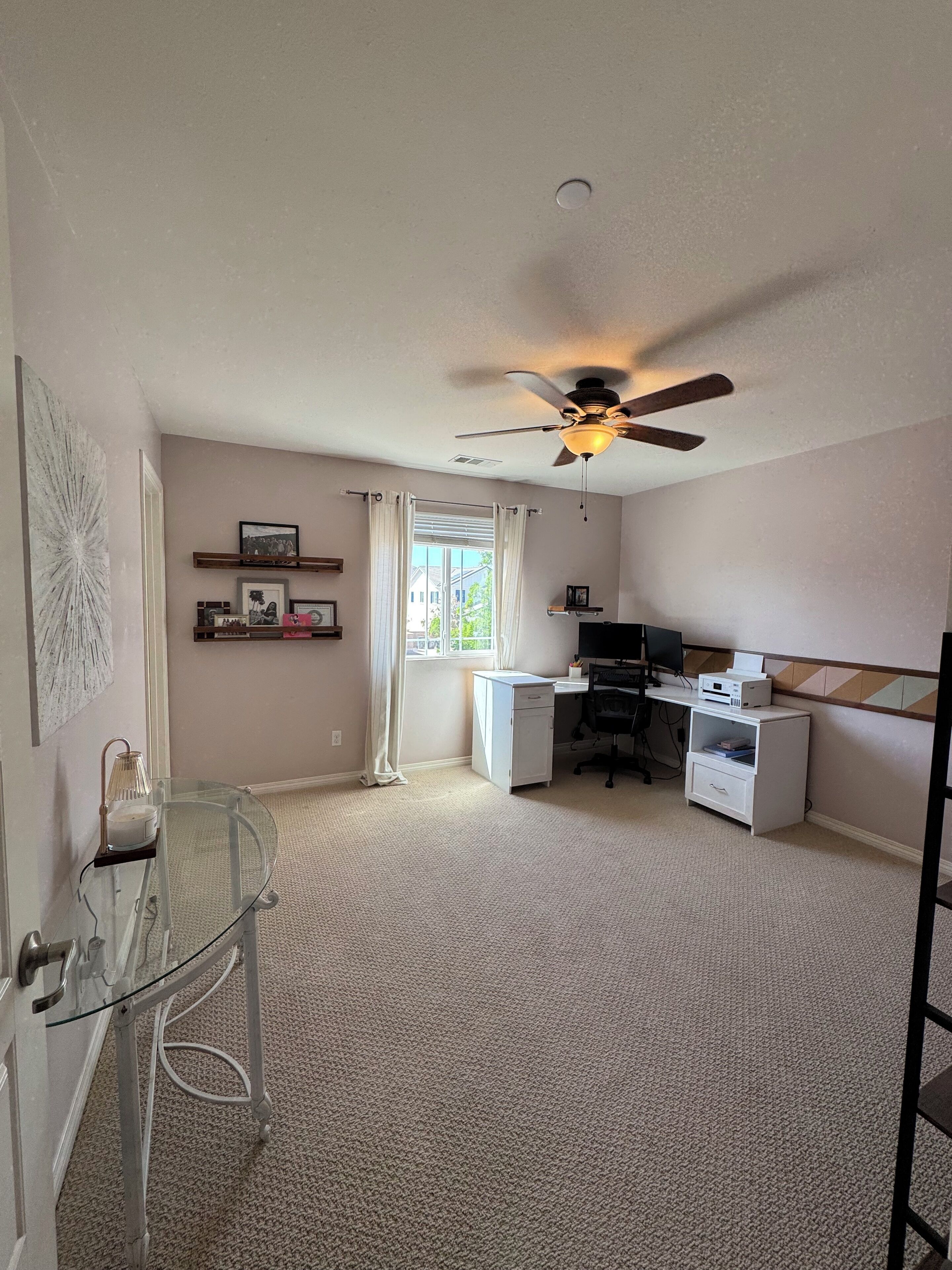 A spacious room with a ceiling fan, a desk with a computer, a printer, and a small cabinet. There is a window with curtains, a door with a glass table, and a wall-mounted shelf with pictures.