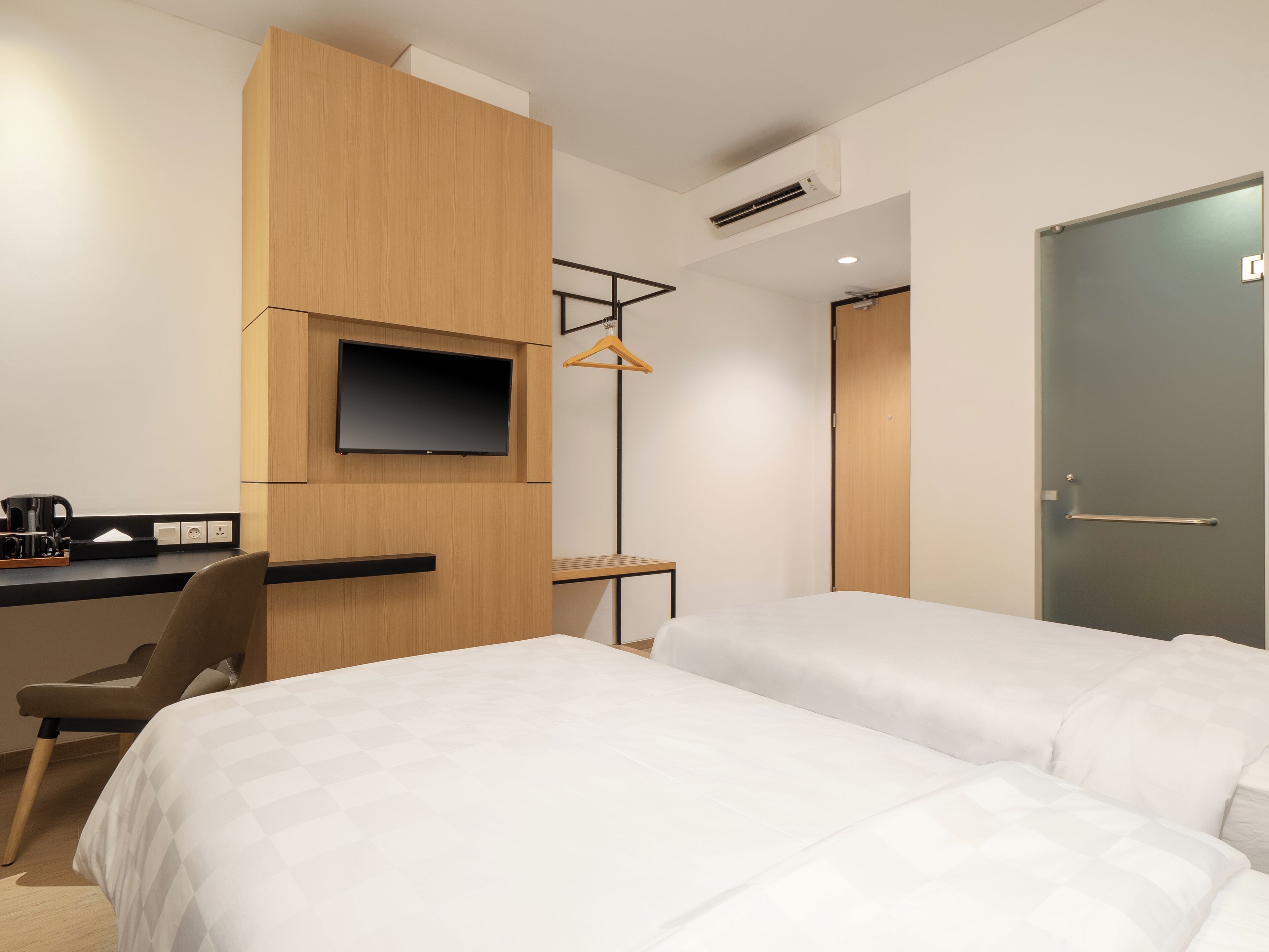 A modern hotel room with two beds, a desk with a flat-screen TV, a coffee maker, and a hanging coat rack.