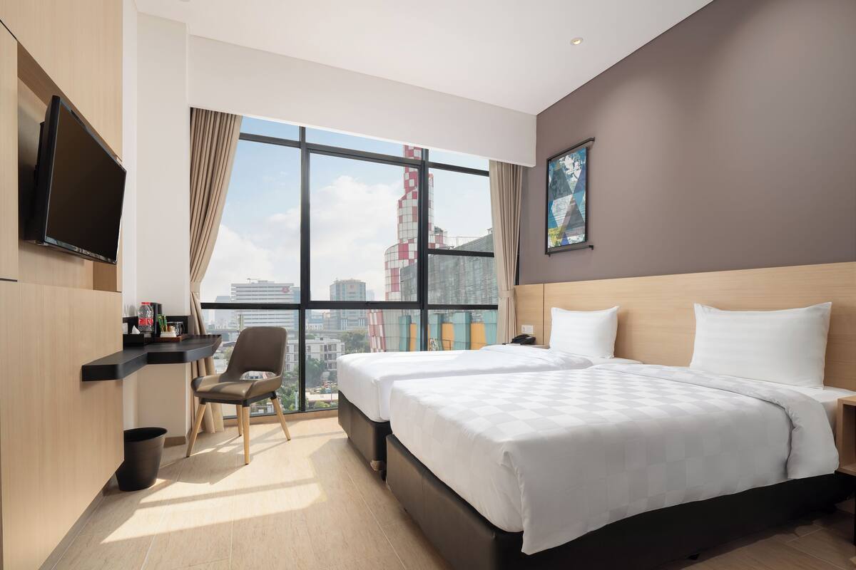 A modern hotel room with a large bed, a TV mounted on the wall, a chair, and a view of the cityscape.
