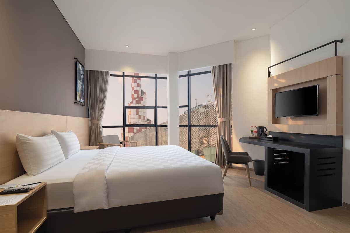 A modern hotel room with a large bed, a desk with a TV, and a city view.