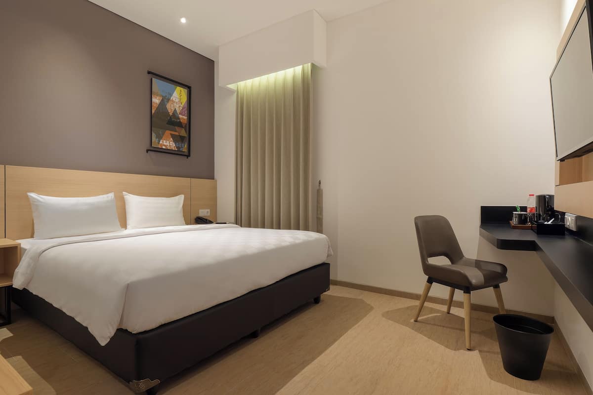 A modern hotel room with a large bed, a desk with a chair, a trash bin, and a painting on the wall.