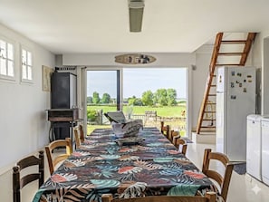 A dining area with a table covered in a floral tablecloth, wooden chairs, a view of the countryside through large windows, a refrigerator, and a ladder leaning against the wall. - Villa Colette by Interhome (Plouhinec Morbihan)