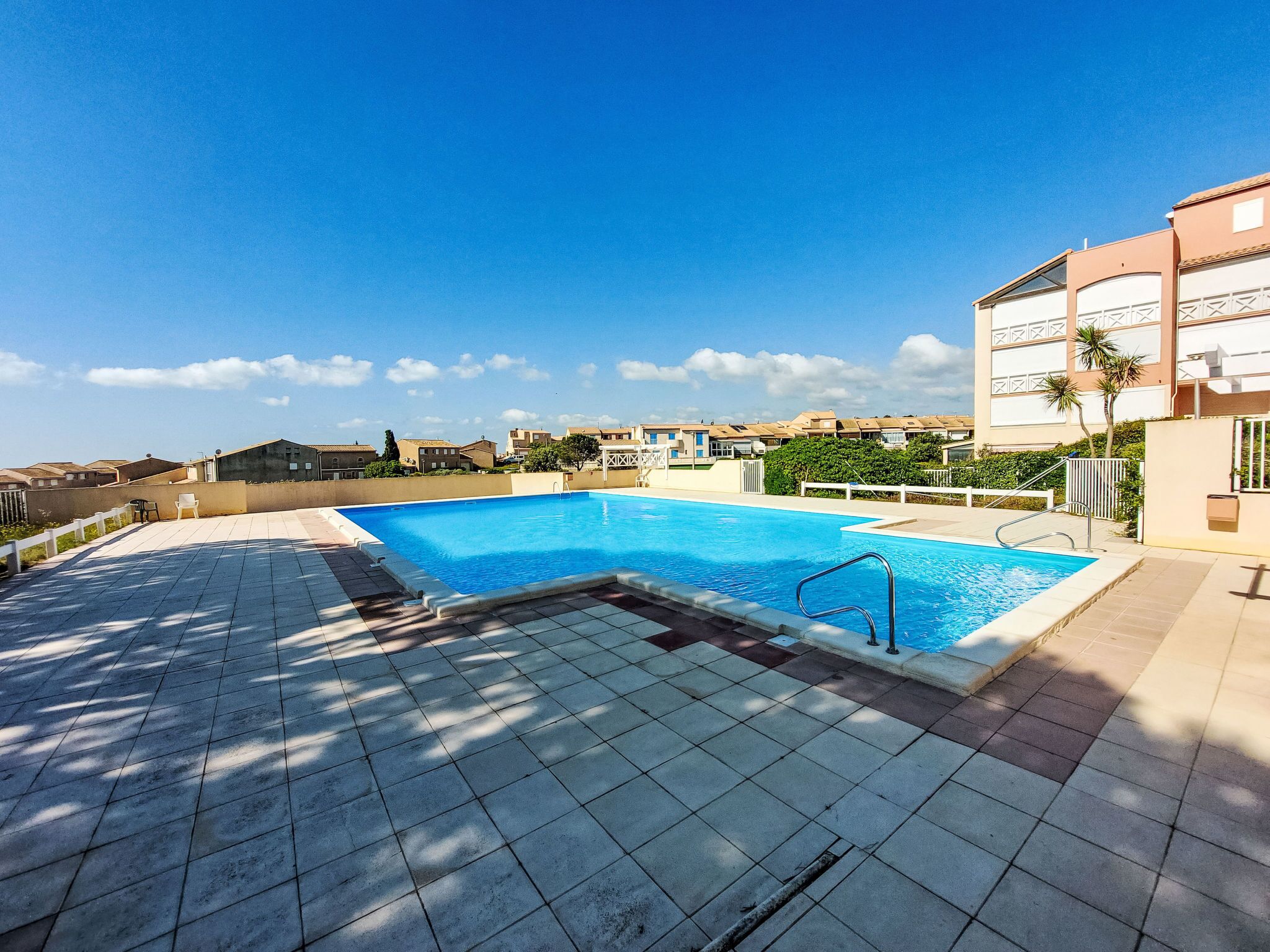 A swimming pool with a tiled deck, surrounded by buildings and greenery.