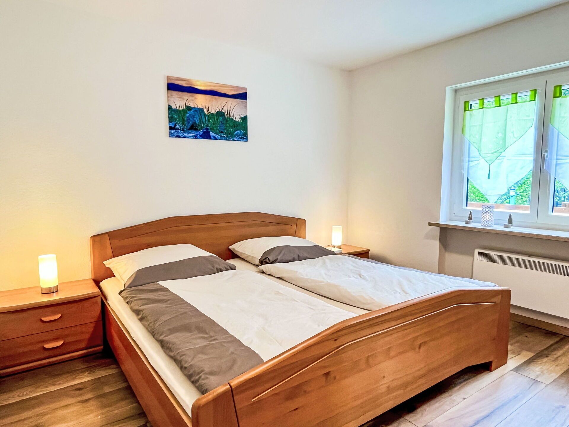 A bedroom with a wooden bed, bedside tables, a nightstand, a window with a view, and a painting on the wall.