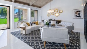 Una sala de estar moderna con un sofá seccional, una mesa de centro redonda y un sillón gris. Hay una araña, decoración en la pared y vistas a un jardín verde a través de amplias ventanas.