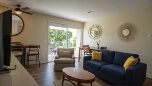 Una sala de estar con un sofá azul, una mesa de centro de madera, una zona de comedor con mesa y sillas, y un balcón con vistas a zonas verdes.