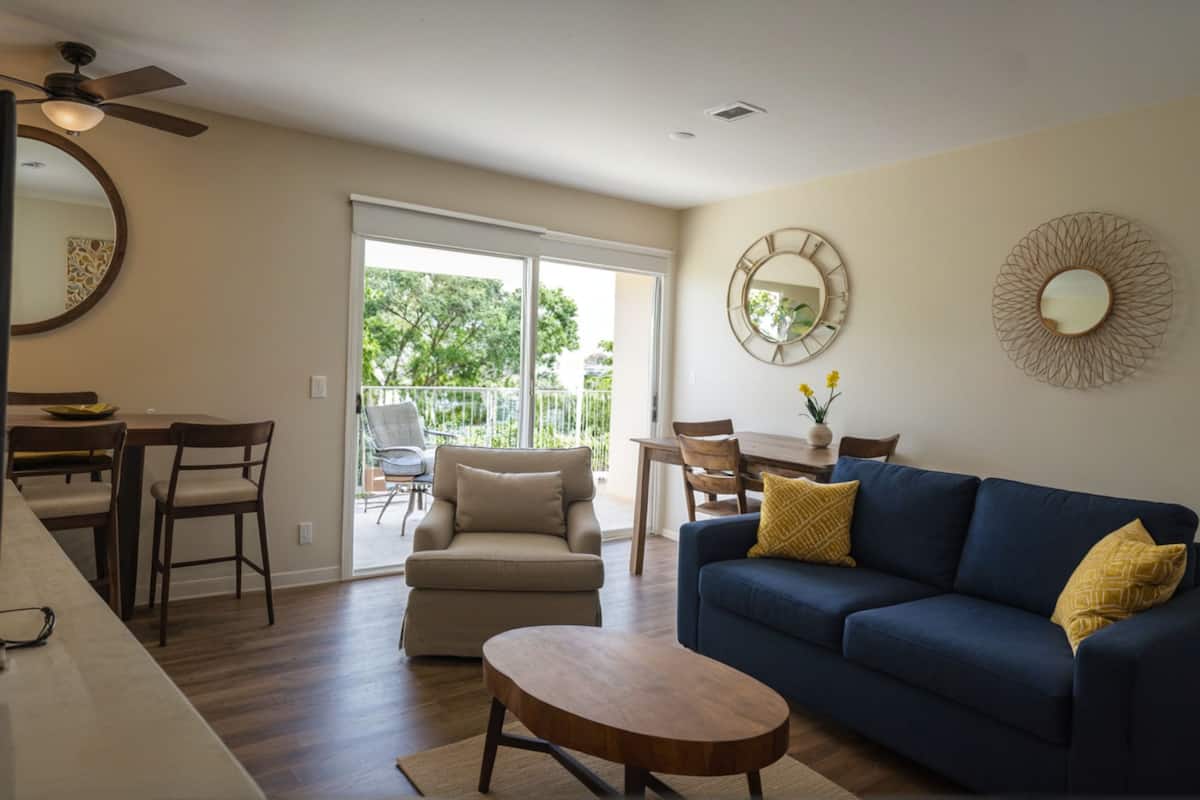 Un soggiorno con un divano blu, un tavolino in legno, un'area pranzo con tavolo e sedie, e un balcone con vista sul verde.
