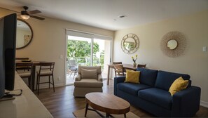 A living room with a blue sofa, a wooden coffee table, a dining area with a table and chairs, and a balcony with a view of greenery. - Executive Suites near Santa Monica (Los Angeles)