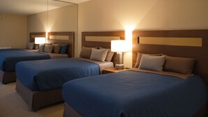 Habitación de hotel con tres camas, cada una con una colcha azul y un cabecero de madera. Hay una mesita de noche con una lámpara entre las camas.