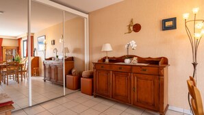 A spacious living room with a wooden cabinet, a dining area with a table and chairs, and a large mirror reflecting the room.