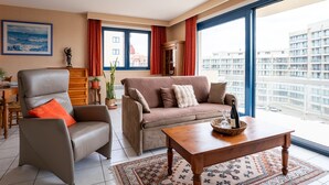 A modern living room with a sofa, armchair, and coffee table, large windows with a city view, and a painting on the wall.