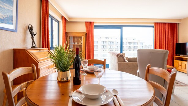 A dining area with a round wooden table, chairs, a bottle of wine, a glass, and a bowl. There is a window with curtains, a television, and a plant on the table.