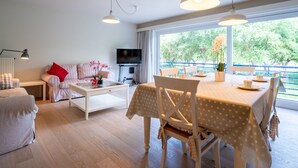 Ein geräumiges Wohnzimmer mit einer Couch, einem Couchtisch und einem Essbereich mit Tisch und Stühlen. Ein Fernseher ist an der Wand montiert, und es gibt ein großes Fenster mit Blick auf Grünflächen.