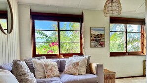 A cozy living room with a sectional sofa, a round wooden side table, and large windows with a view of greenery. - Cosy Manly Beach Hideaway (Manly)