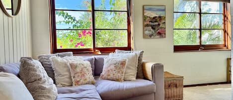 A cozy living room with a sectional sofa, a round wooden side table, and large windows with a view of greenery.