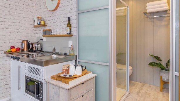 A compact kitchen with a sink, stove, and microwave, a wooden countertop with fruits and a kettle, and a glass shower with a towel rack.