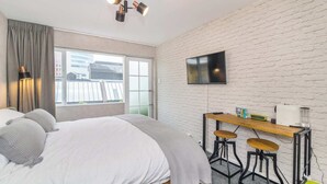 A modern hotel room with a large bed, a desk with a chair, a TV mounted on the wall, and a window with a view of buildings.