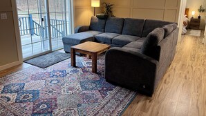 A living room with a sectional sofa, a small wooden coffee table, a patterned rug, and a sliding glass door leading to an outdoor deck. - Whispering River / EV / Mins to Windham and Hunter Mtn (Hensonville)
