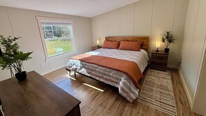 A bedroom with a wooden bed, a nightstand, a bedside table, a window, and a potted plant. - Whispering River / EV / Mins to Windham and Hunter Mtn (Hensonville)