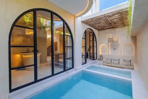 A modern indoor pool area with clear glass walls, a seating area with cushions, and a ceiling with hanging lights. - Villa Santa Clara by Pertama Stay (Seminyak)